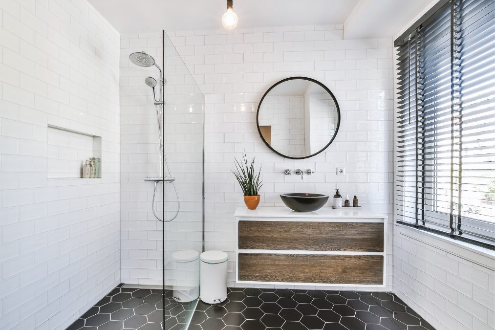 A modern, clean bathroom with white subway tiles, a walk-in shower with a glass door, a round mirror above a sink, and a wooden vanity against a black hexagonal tiled floor. MK plumbing & Heating (1)