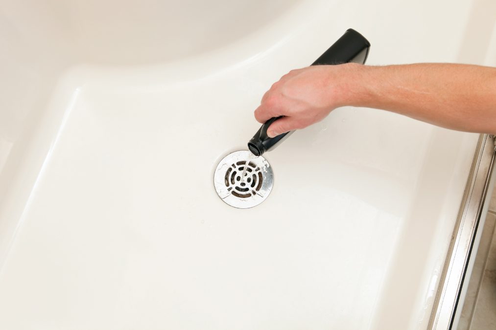 person using a black chemicals to unclog a white bathtub drain with a silver cover. MK plumbing & Heating dorset pluming